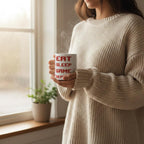 Woman in oversized cream chunky knit sweater holding steaming "Eat Sleep Game Repeat" gaming mug by sunlit window with small houseplant – cozy gamer aesthetic gift mug